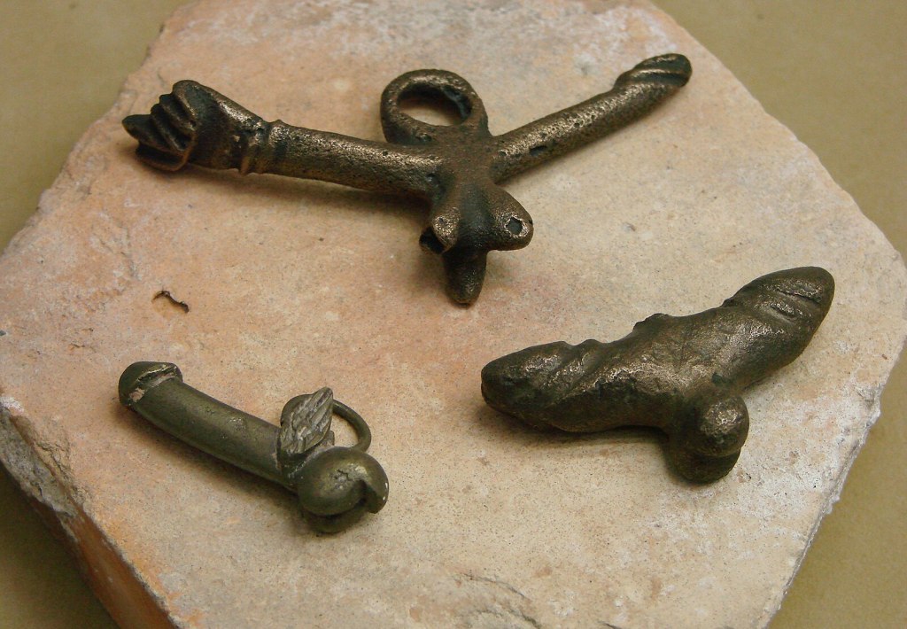 Three phallic amulets made of metal, as displayed at a museum in Reims, France.