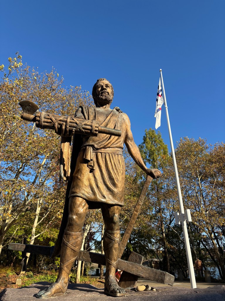 A photograph looking up at a large bronze-colored statue of the Roman statesman Cincinnatus in front of a flag of the city of Cincinnati. He is holding up the fasces in his right hand as he leans on an plow with his left.