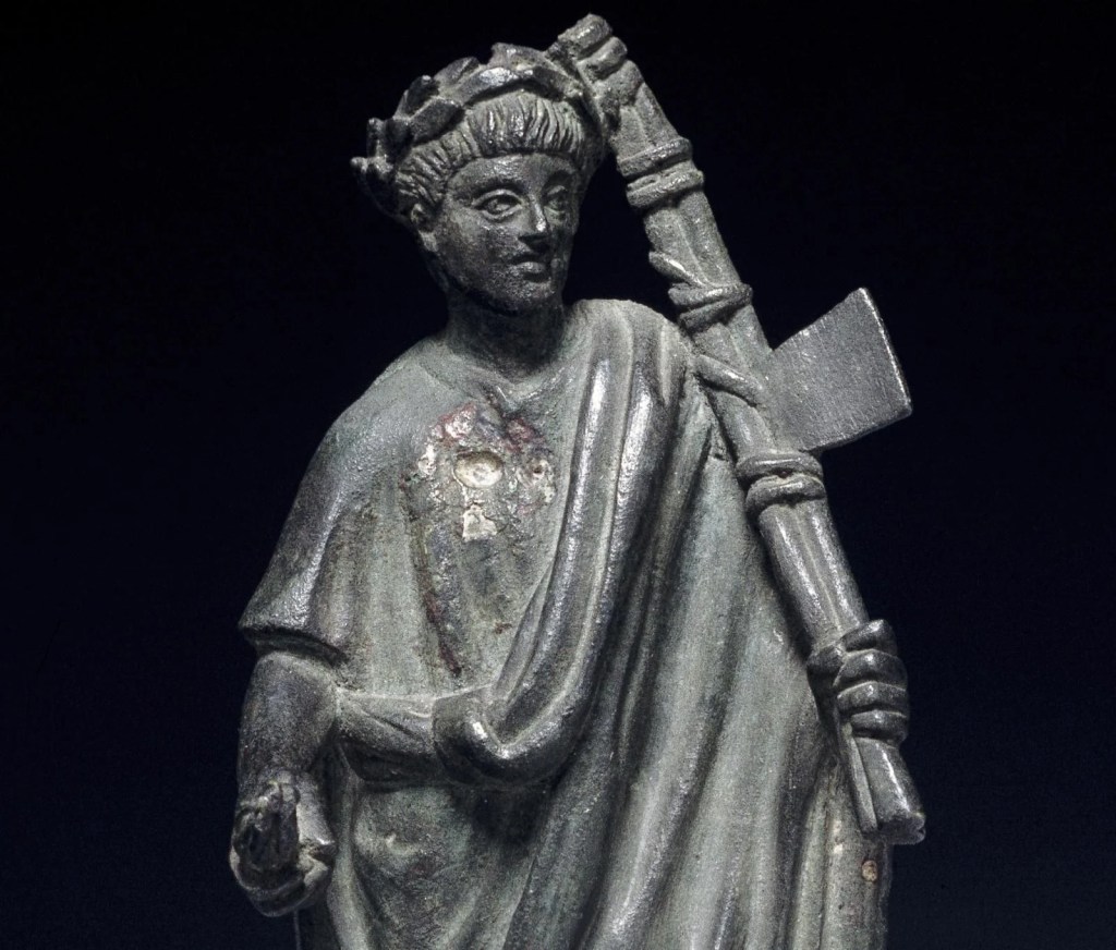 A photograph of a small statue from Ancient Rome showing a lictor (attendant) bearing the fasces wearing a toga and a wreath on his head.