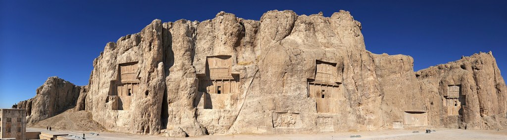 A panorama of the Naqsh-e Rostam compound in Iran showing the Kaaba-ye Zartosht on the far left and arraying the rock reliefs of the tombs of Achaemenid kings.
